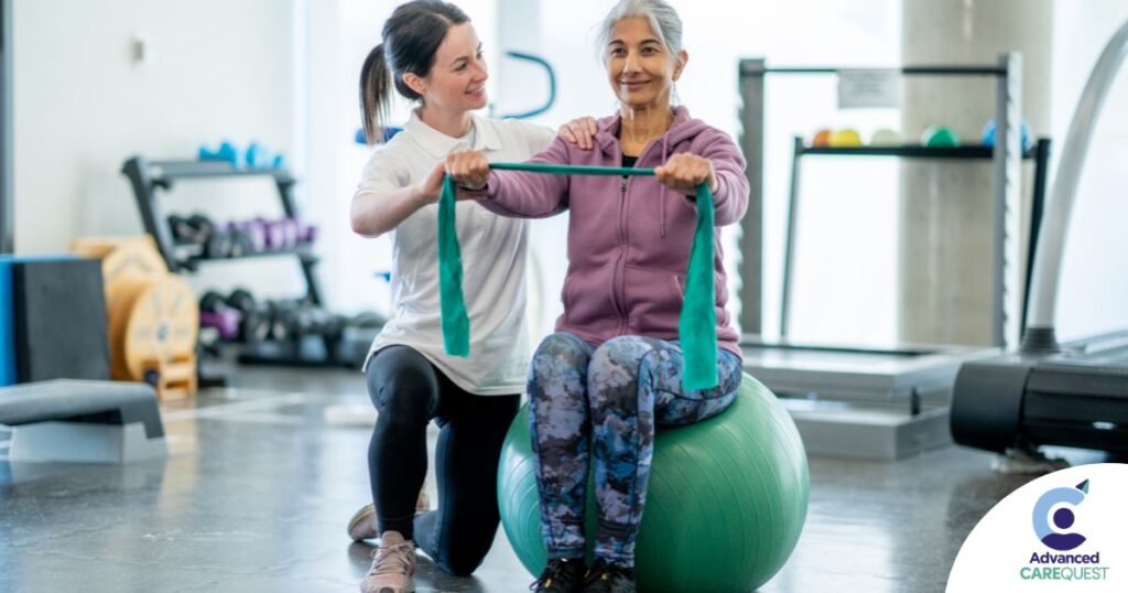 A care provider helps an older woman exercise with a resistance band and an exercise ball, representing how exercise can help with senior fall prevention.