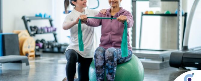 A care provider helps an older woman exercise with a resistance band and an exercise ball, representing how exercise can help with senior fall prevention.
